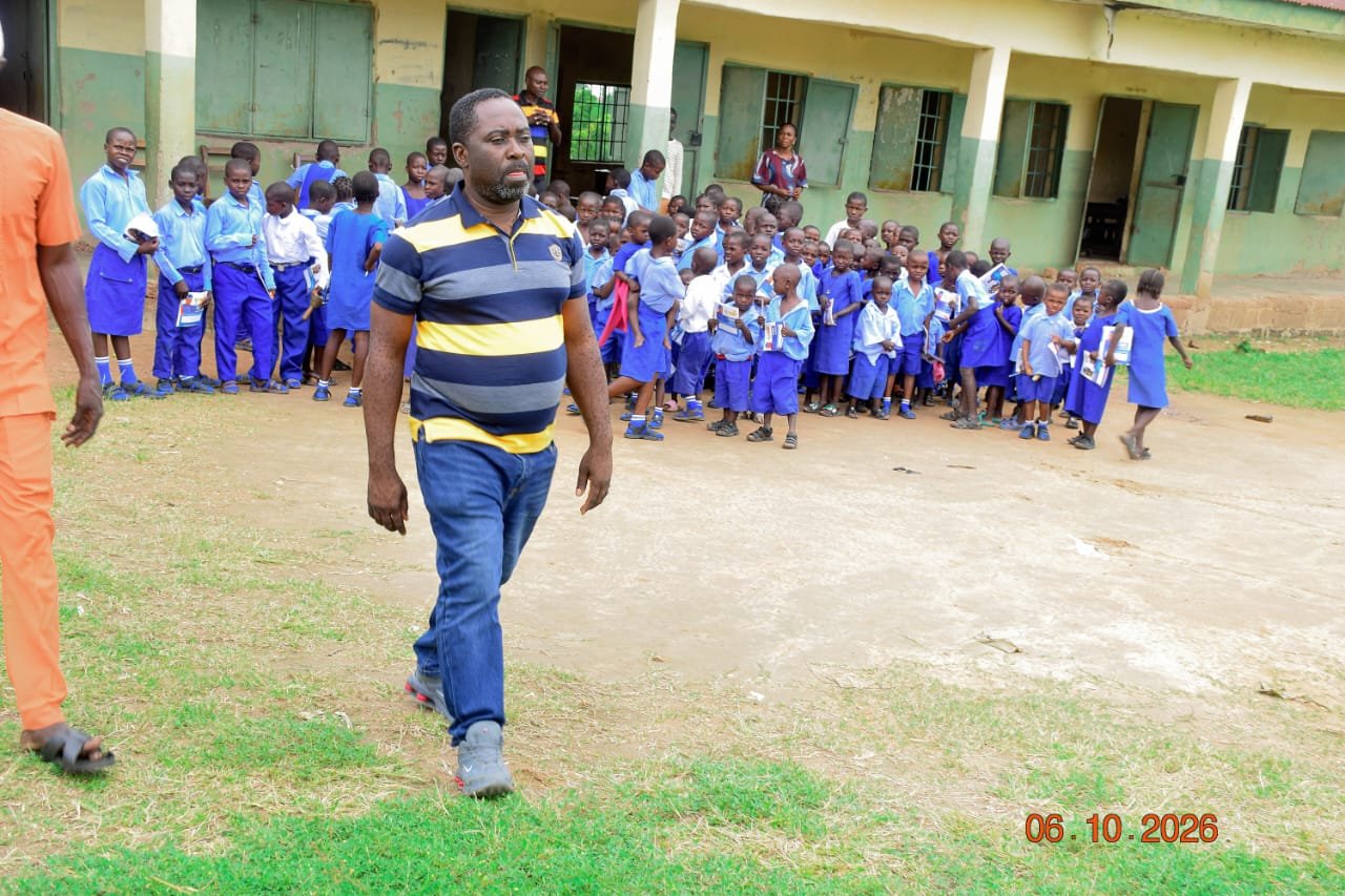Agber walking across a grassy school compound with pupils gathered in front of the classroom block