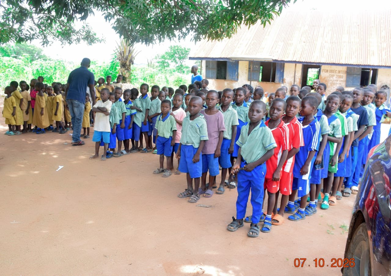 Pupils in green checked uniforms lined up under a mango tree beside an FGN UBEC wall