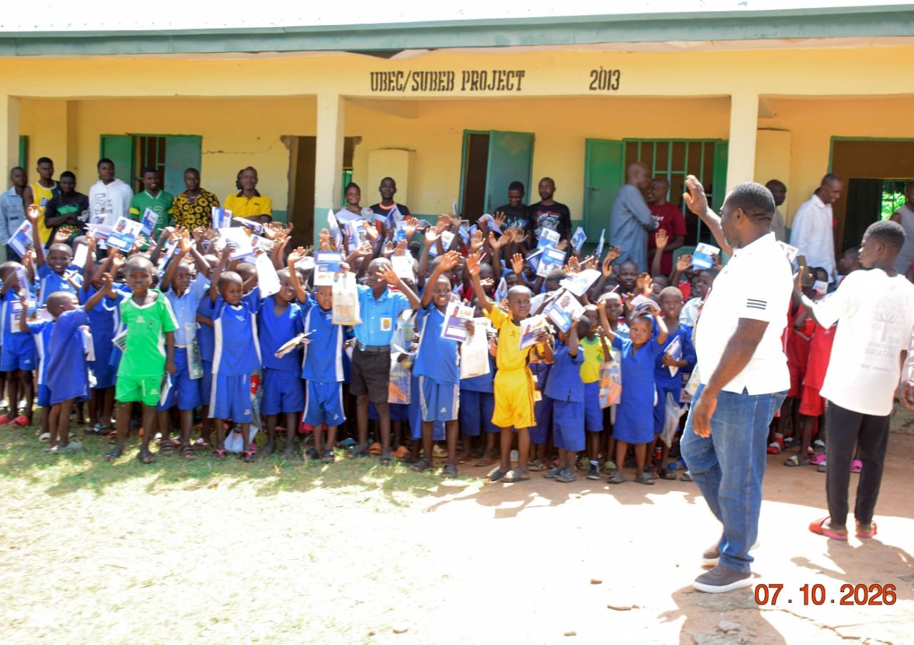 Agber in a white polo with his hand raised before pupils at a yellow UBEC/SUBEB school