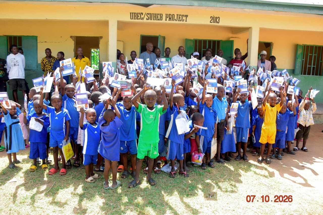 Group photo of pupils raising branded exercise books against the yellow walls of a UBEC/SUBEB school