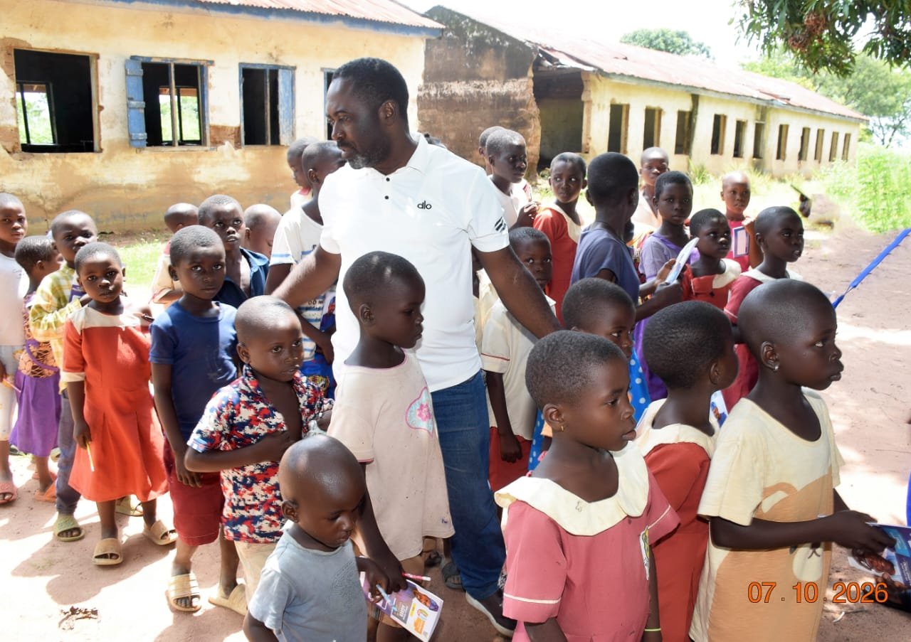 Agber in a white polo standing on red earth beside a long line of pupils at a green roofed school
