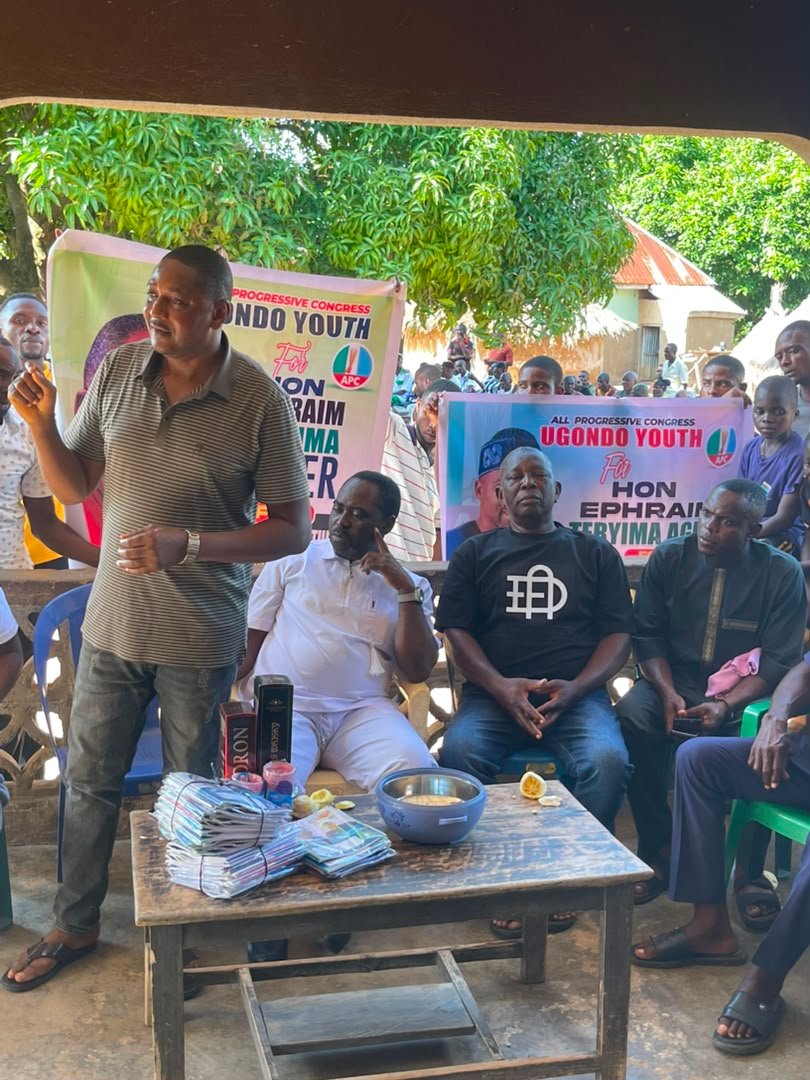 A supporter in a yellow shirt addressing the Ugondo community meeting beneath APC banners