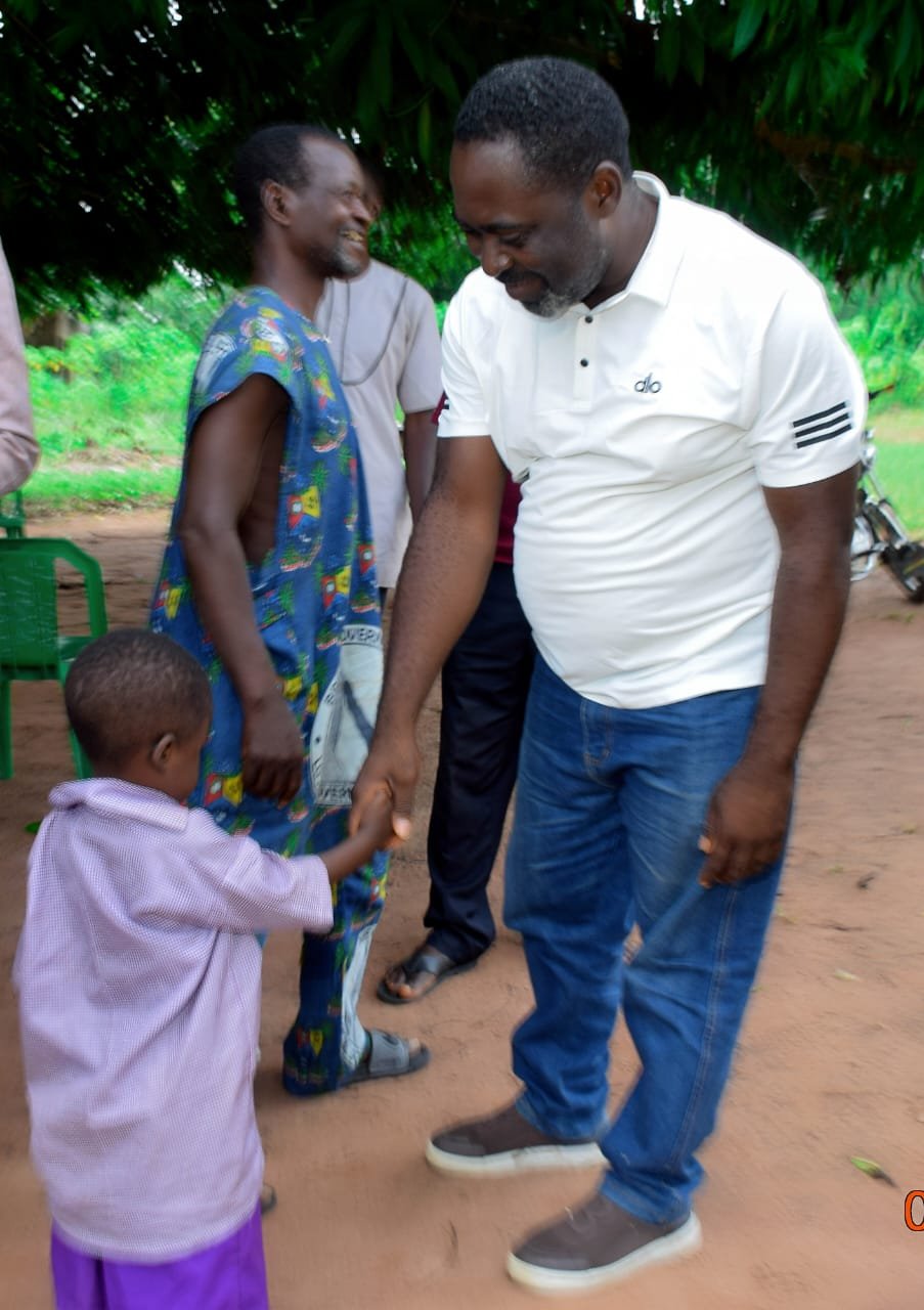 Agber in a white polo shaking hands with a small pupil while an elder in blue smiles beside them
